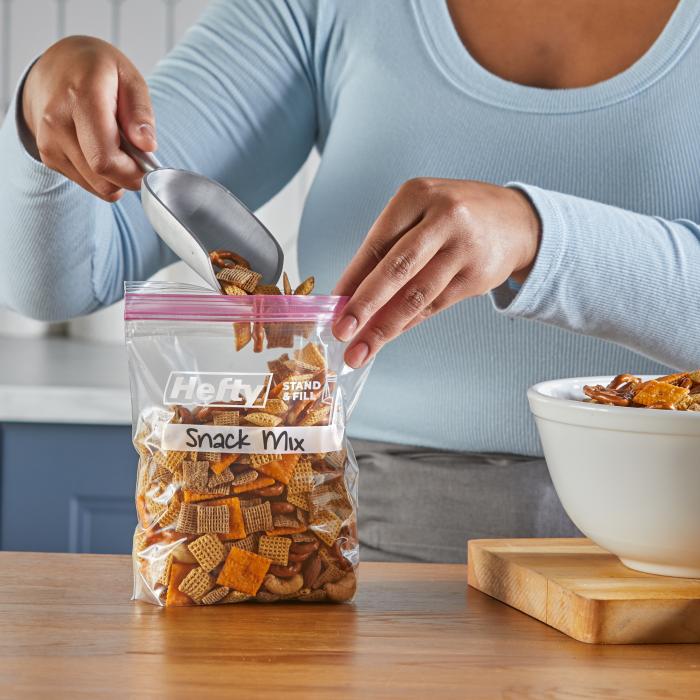 A person filling a quart sized Hefty Press to Close bag with snack mix using a scoop