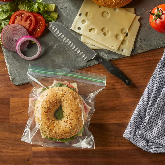 Bagel sandwich in a sealed Hefty Press to Close Sandwich XL sized storage bag alongside a cutting board with sandwich ingredients 