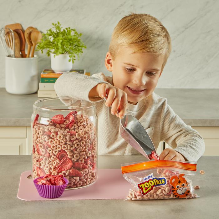 child using a scoop to pour strawberry cereal trail mix into a Hefty Zoo Pals snack size storage bag